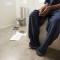 Juvenile solitary: Close-up of legs and hands-in-lap of black person wearing over-sized navy pants sitting on edge of built-in cement bench with metal toilet in background.