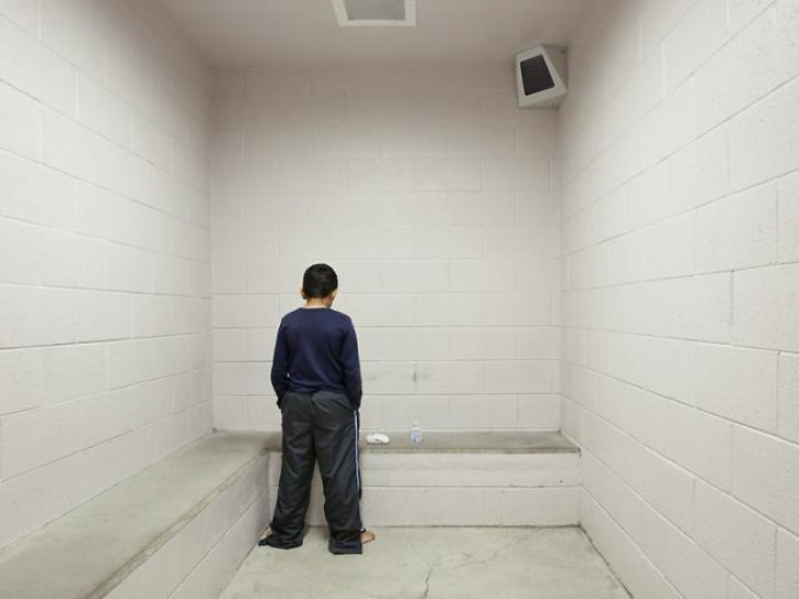 Juvenile solitary: Youth in all dark shirt & pants tansa weth back to camera in wdire cell wit built-in cement bed