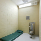 Juvenile solitary: Small, brightly lit room with no windows and a metal wall urinal next to a green plastic sleeping pad in the corner on the floor.