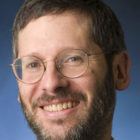 Daniel Pollack (headshot), Yeshiva University professor; smiling man with beard, mustache, glasses wearing blue tie, white shirt, dark vest. Feature photo alt text: Blank-faced child in big shirt, pants stands against height chart.