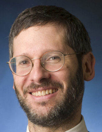 Daniel Pollack (headshot), Yeshiva University professor; smiling man with beard, mustache, glasses wearing blue tie, white shirt, dark vest. Feature photo alt text: Blank-faced child in big shirt, pants stands against height chart.