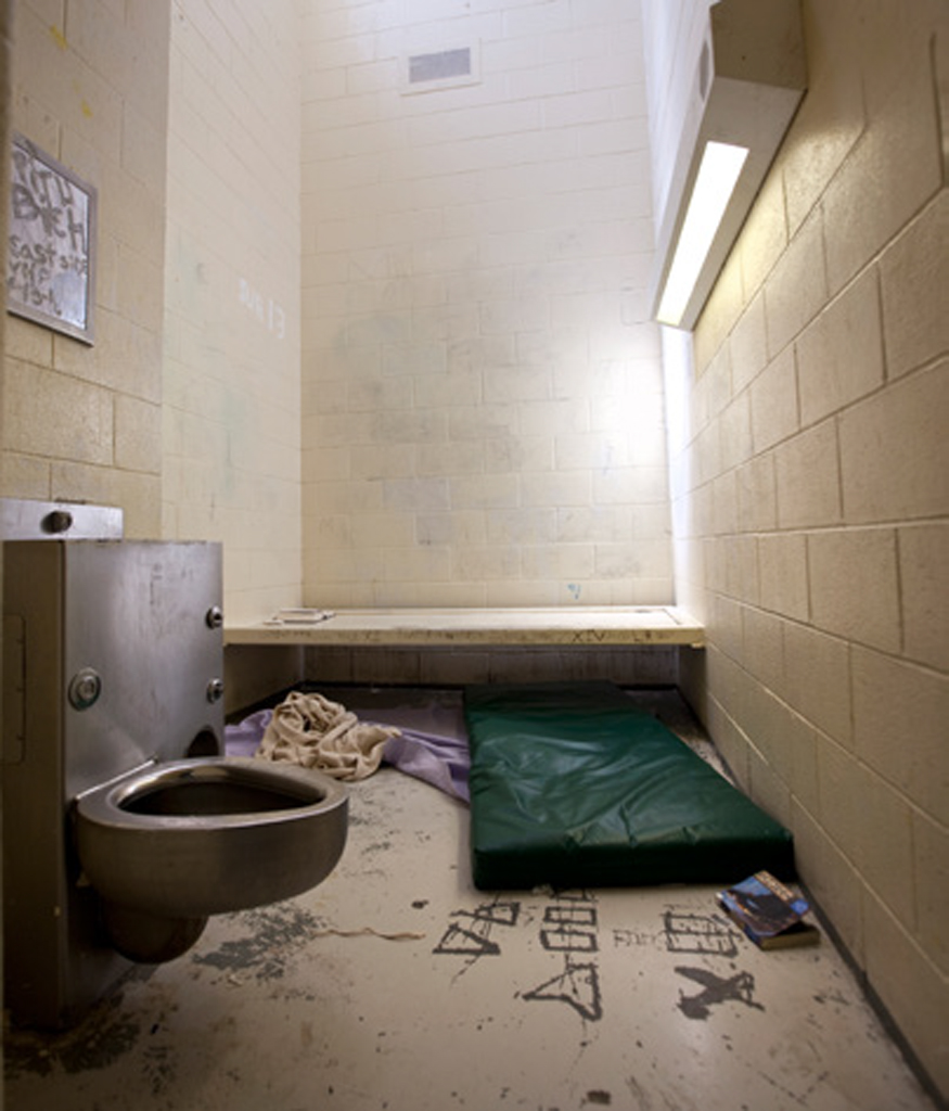 Juvenile solitary: Small cell with green vinyl sleeping pad and blankets on floor, metal toilet and florescent light attached to wall, and graffiti scraped through paint on floor.