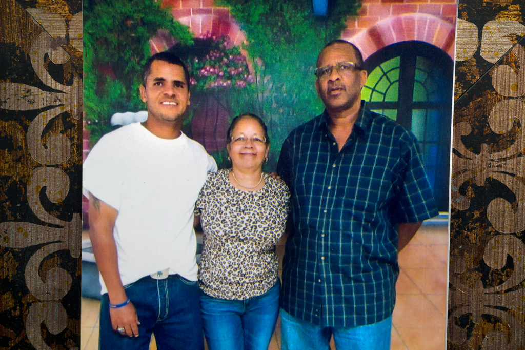 Close-up of another photo displayed: Smiling young man in white T-shirt, blue jeans, big belt buckle, blue rubber bracelet on right hand posing with shorter woman on his left. She's wearing leopard-print short-sleeved top, blue jeans, glasses and is smiling. To her left is older man, unsmiling, with glasses, blue and green checked untucked shirt and blue jeans.