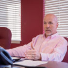 White man in pink shirt with gray hair, mustache and beard sits at desk facing to left of center and gestures as he speaks.