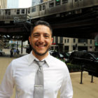 David Blancas, in a white shirt and gray tie, smiles as he stands outside his Chicago office, an elevated train above him.