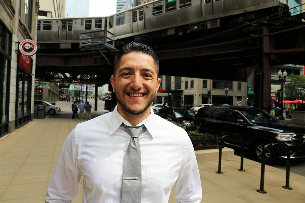 David Blancas, in a white shirt and gray tie, smiles as he stands outside his Chicago office, an elevated train above him.