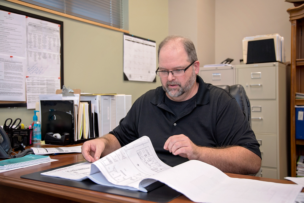 Balding man with greying short beard looks over papers at his desk.