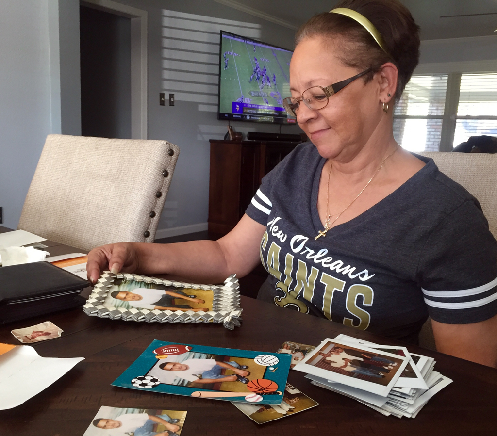 Woman wearing short-sleeved V-neck New Orleans Saints T-shirt, with glasses, hairband, cross on a necklace, small earrings looks down at desk with several photos of same young man.
