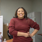 Woman in orange sweater, large hoop earrings smiles widely in office kitchen.