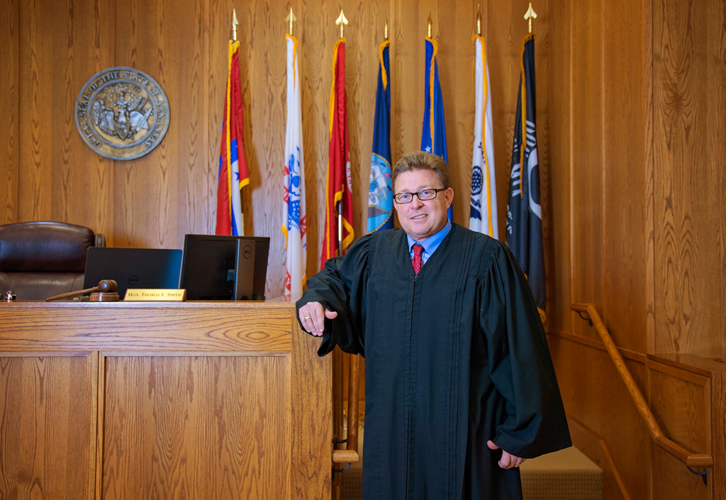 Judge Tom Smith, in his judicial robes over blue shirt and red tie, leans on his bench in the courtroom.