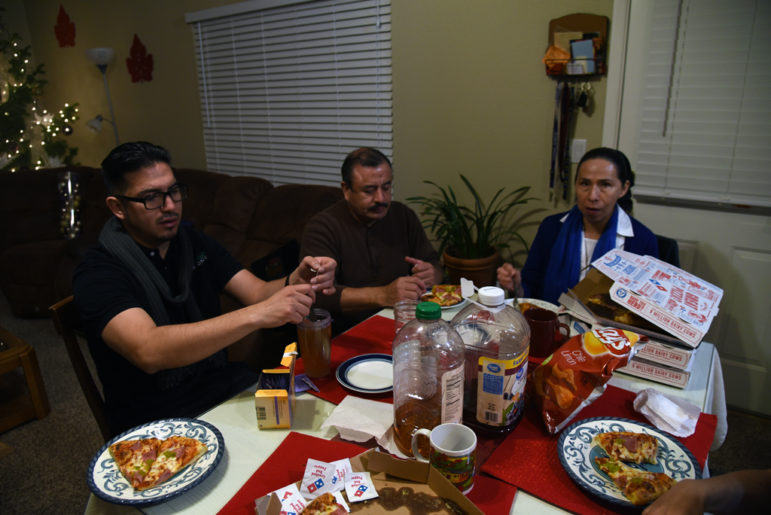 Man with black-framed glasses sits with older couple at table laden with takeout pizza, jugs of fruit juice and a bag of potato chips.