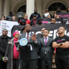 Man in gray suit speaks at outside dais, flanked by people in puffy coats, one man in T-shirt that says Close Rikers and banners with Close Rikers and The Legal Aid Society on them.