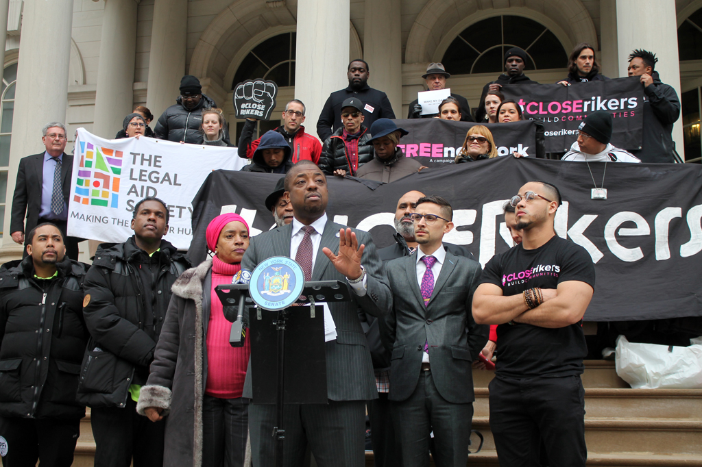 Man in gray suit speaks at outside dais, flanked by people in puffy coats, one man in T-shirt that says Close Rikers and banners with Close Rikers and The Legal Aid Society on them.