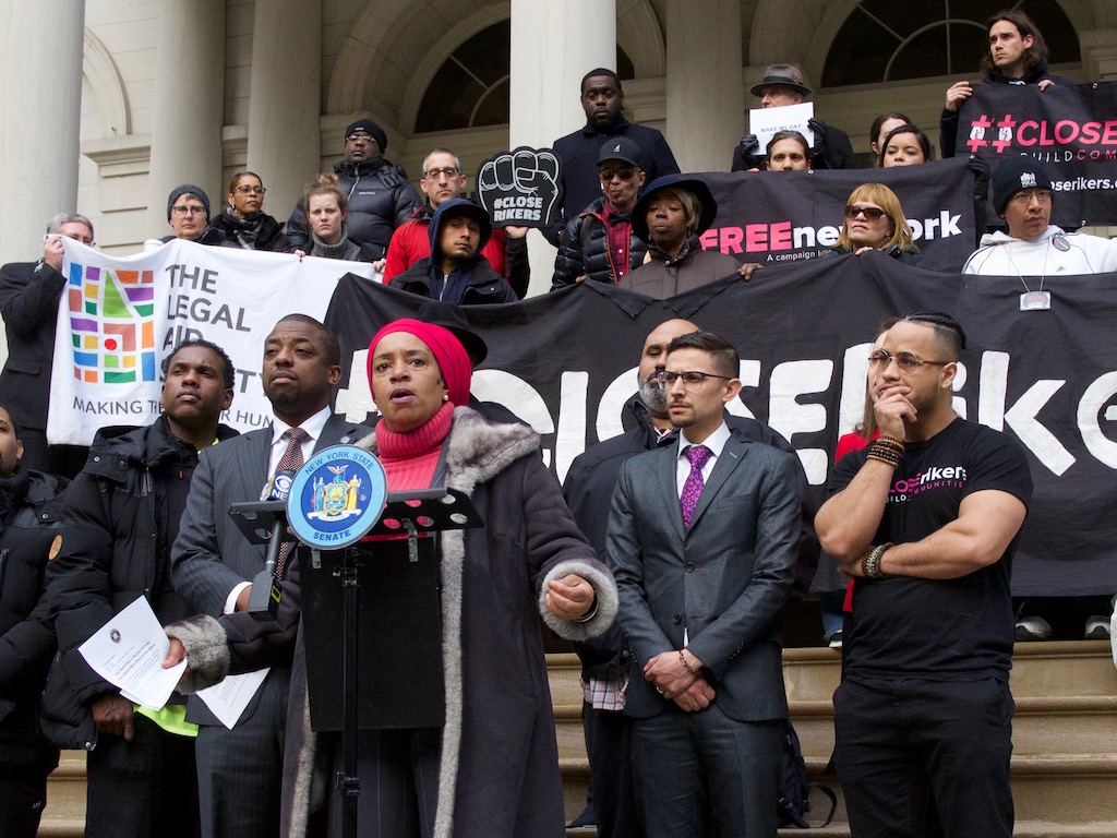 reentry, trauma, Rikers Woman in pink hat, turtleneck speaks on steps of government building, surrounded by men in dark suits, T-shirts, holding banners.