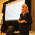 OJJDP Head Caren Harp, wearing dark suit and earrings, holds microphone and stands by flip board with video screen behind her.