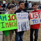 March for Our Lives: 3 boys hold up their signs; one in glasses and tan pants; one in dark jacket and jeans; one in red jacket and dark jeans.