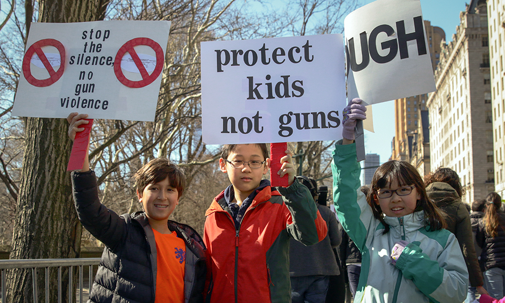 March for Our Lives: 3 young people in winter jackets, a girl in black, a boy in orange and green and a girl in light and dark green, hold up signs for gun control.