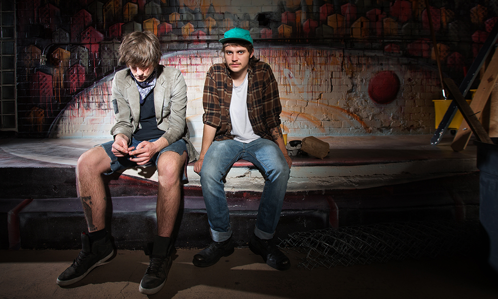 Probation: 2 young men sit on bench against wall; one wears denim shorts, black shoes, olive jacket and looks down; the other wears cuffed jeans, dark shoes, white T-shirt under open brown flannel shirt and green cap, has mustache.