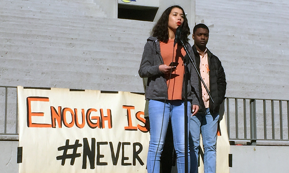Student activist Nowell Mendoza spoke on a stage with large sign behind her, another student to her left.