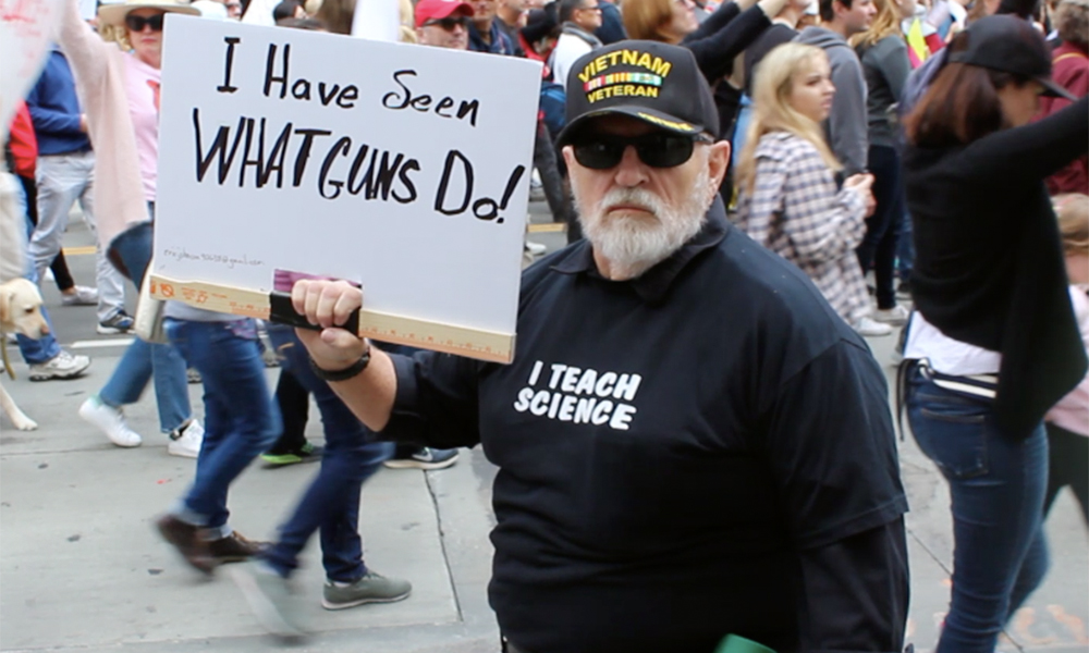 March for Our Lives: A protestor with a gray beard and mustache and dark sunglasses, wearing a dark sweatshirt that says, "I teach science" and a dark cap labelled "Vietname Veteran," carries a sign that says, "I have seen what guns do."