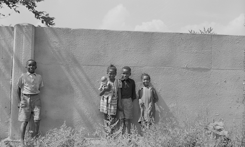 Segregation: 4 smiling children of color stand smiling against wall twice their height in 1941 black and white photo.