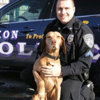 Dixon Police Detective Jeff Ragan (headshot), smiling man in police uniform kneeling in front of police car with left hand on dog’s chest.