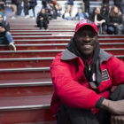 Rikers Island: Smiling man in red work jacket, matching red cap sits on red stairs of Times Square.