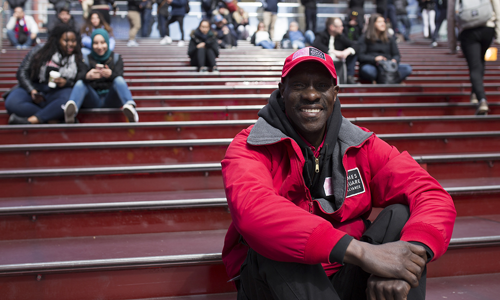 Rikers Island: Smiling man in red work jacket, matching red cap sits on red stairs of Times Square.