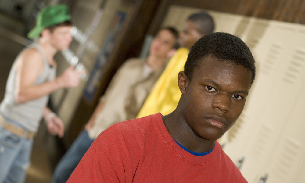 DMC: Serious-looking African-American teen in red T-shirt stands alone near locker, 3 other teens are in background.