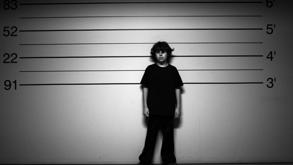 Blank-faced child in big shirt, pants stands against height chart.