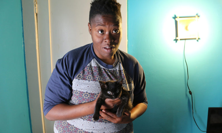 Homelessness: Woman holds bug-eyed, tiny black kitten; bright light on wall on right.