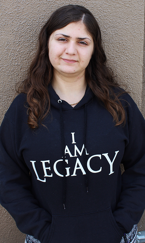 Young woman with long brown hair stands against wall, wearing dark blue hoodie that says I am legacy.