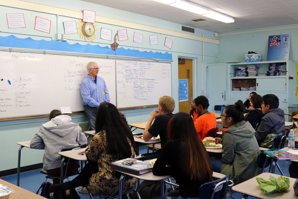 POPS: Man with white hair, glasses, light blue shirt standing against whiteboard talks to high school students sitting at individual desks.