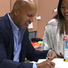 Bryan Stevenson: Smiling bald man in light blue shirt, dark blue jacket signs book for smiling woman with long dark hair in white jacket, dress, with ID badge around her neck.