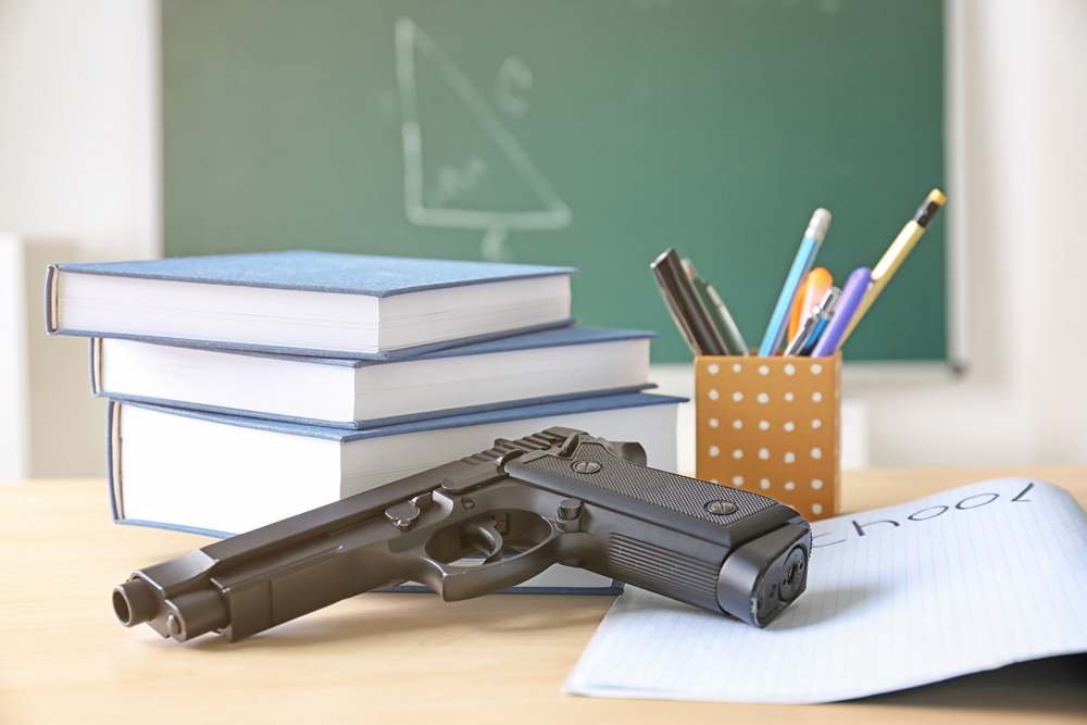 Gun violence: Gun lying on desk with 3 blue-covered books, paper with word School on it and pencil holder with pens, pencils, ruler; geometry drawing on blackboard is in background.