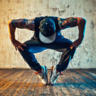Young tattooed man in ball cap break dancing, on his toes, hands on knees, on wall background.
