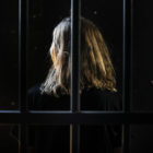 Woman seen from back, dressed in black, behind bars, with only hair spotlighted.