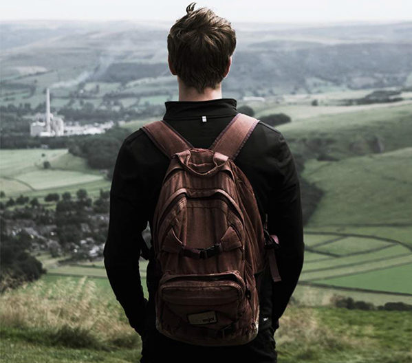 Homelessness: Young man wearing backpack stands at top of hill gazing down at green vista.