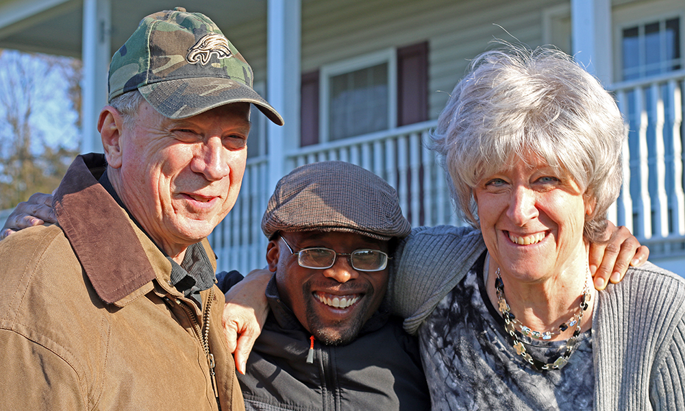 Older man in jacket wearing camouflage cap smiles on left. Next to him is young man in jacket, wireframe glasses, newsboy cap and older woman with gray hair, necklace, cardigan over print top; they have arms around each other. 