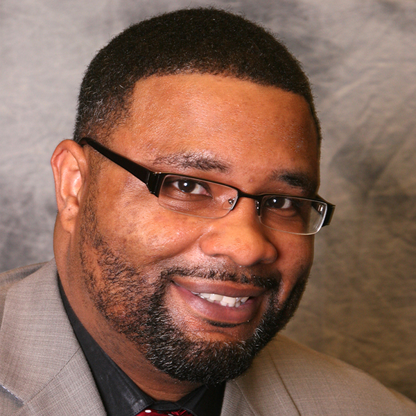 Solitary confinement: DeShane Reed (headshot), senior juvenile justice practitioner with DRB Consulting, smiling man with mustache, beard, wearing beige jacket, dark shirt, red tie with black and white polka dots.