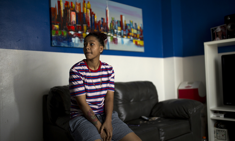 Trauma: Young woman in jean shorts, striped T-shirt, necklace, tattoos, looks to side on couch in front of bright painting.