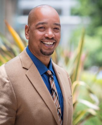 Tshaka Barrows (headshot), Executive Director of W. Haywood Burns Institute, smiling man in blue polka dot shirt, patterned brown tie, beige jacket
