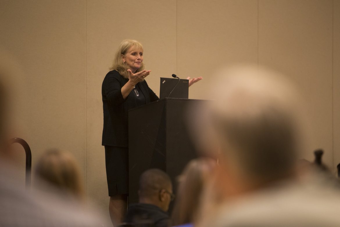 DMC: Blonde woman in black outfit at podium gestures.