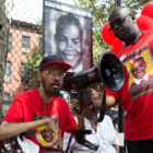 police shootings: Haggard man sits on walker speaking through megaphone surrounded by man and woman; all 3 wearing matching red T-shirts with boy’s stylized illustration on it.