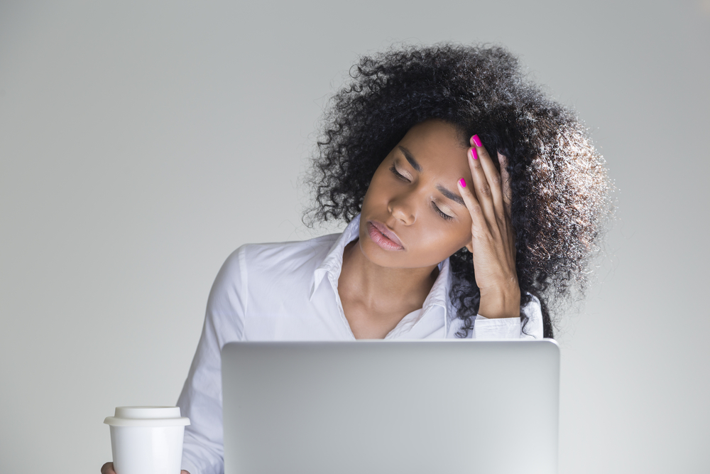 crisis planning: Closeup of tired office employee sitting at her workplace, head in hands, with laptop in room with gray walls.