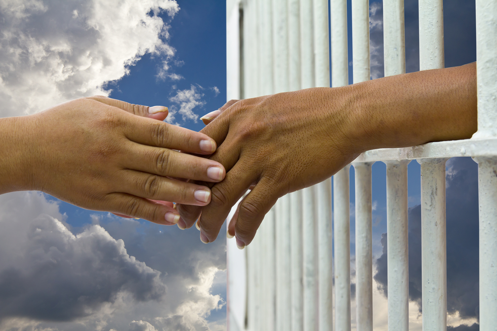 Woman’s hand touches hand of husband to prison with a cloud sky background. Tags: freedom, love, sacrifice