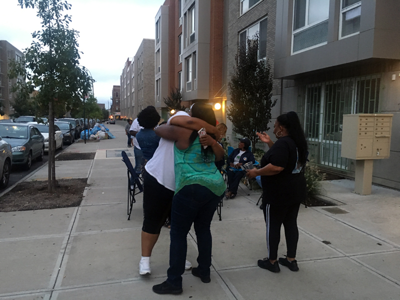 murderversary: 2 people hug on sidewalk, others sit on folding chairs nearby.