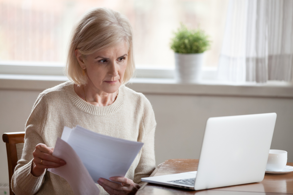 Serious aged woman holding documents, checking information at laptop online