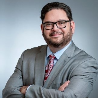New York City: Felipe Franco (headshot), deputy commissioner of New York City Division of Youth and Family Justice, smiling man with glasses, beard, mustache, arms folded in gray suit, light blue shirt, maroon paisley tie
