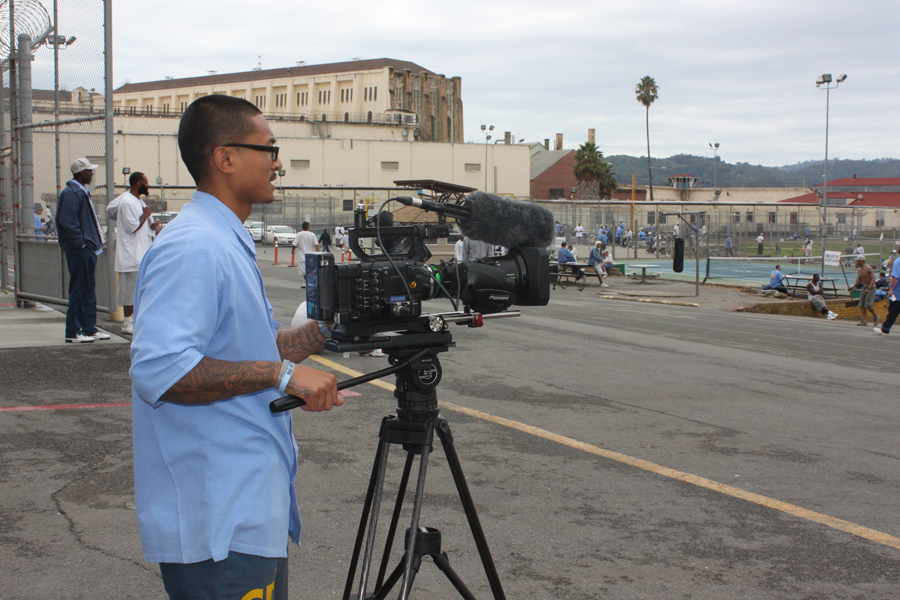 San Quentin: Man in light blue, glasses stands at camera in institutional yard.